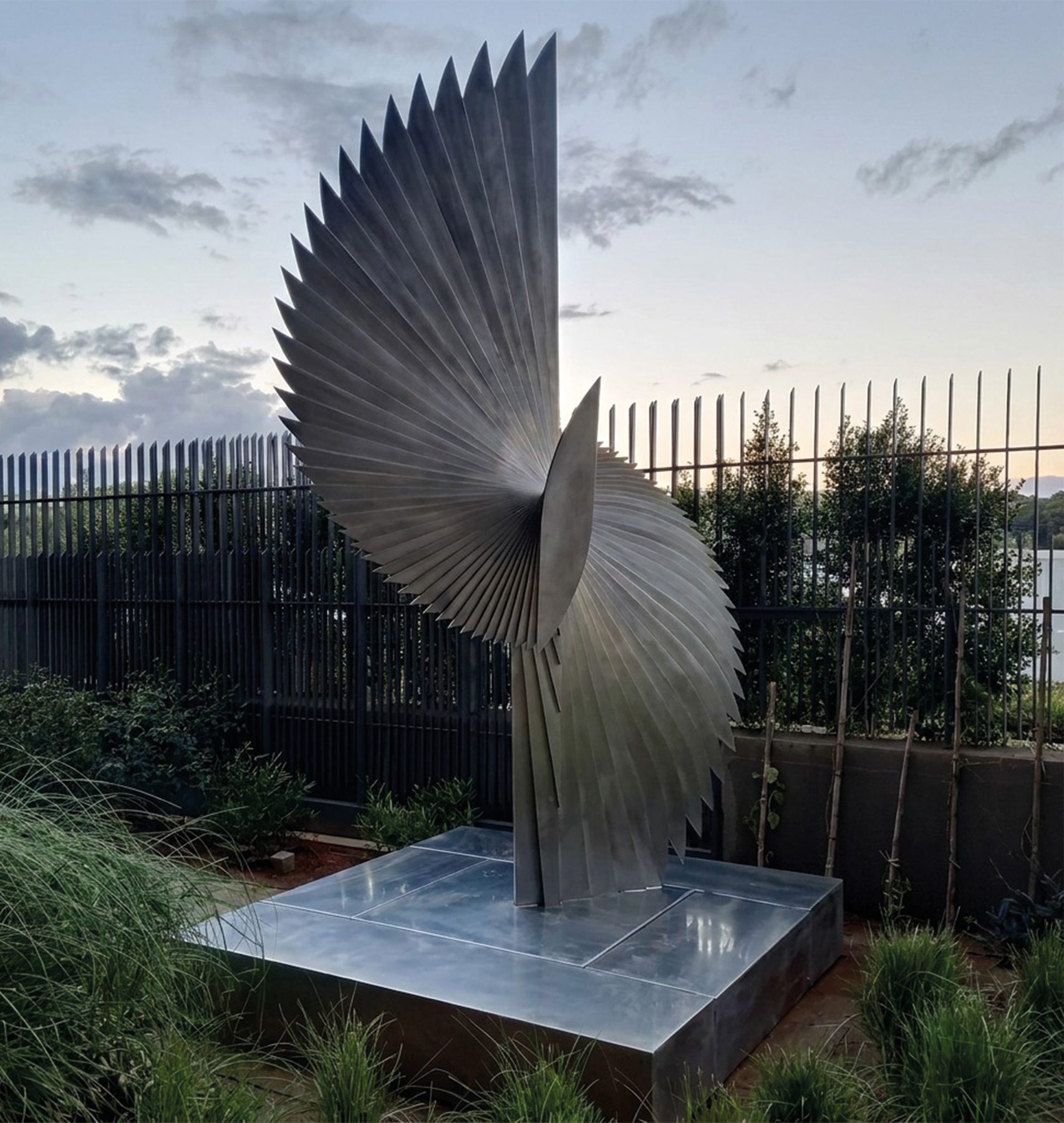 Sculpture monumentale en forme d'aile déployée en acier inoxydable lamellé, installée sur socle dans un jardin au crépuscule, œuvre d'art extérieure sur mesure réalisée au Maroc
